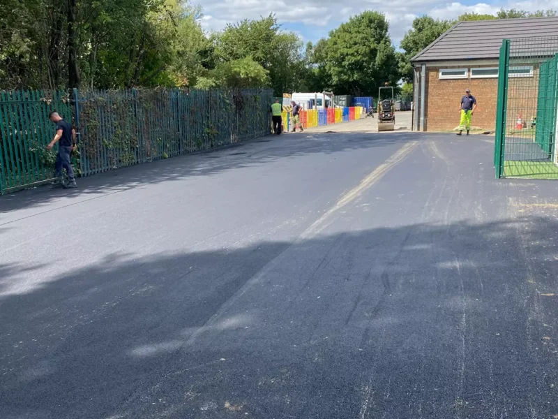 Team working on large tarmac area in front of a brick building and green metal fence.